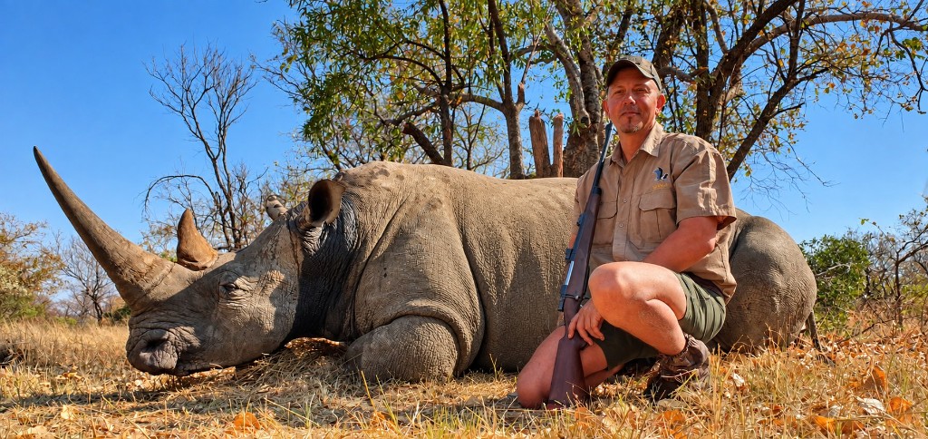 White rhino lying in dry grass with a kneeling hunter in khaki holding a rifle under a blue sky